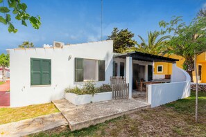 Un pequeño edificio blanco con contraventanas verdes, un patio y un edificio amarillo al fondo.