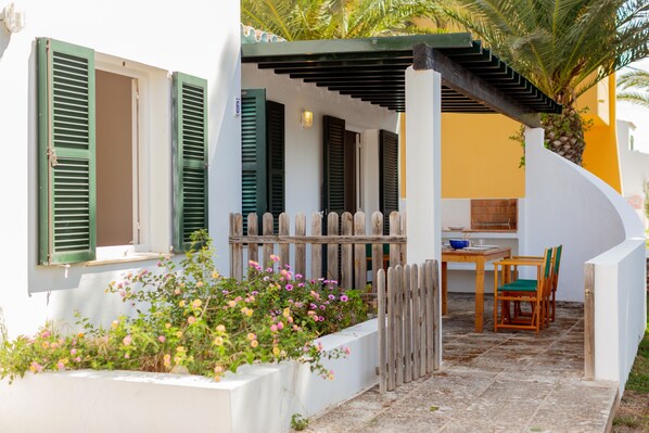 A white building with green shutters, a wooden fence, and a patio area with a table and chairs.