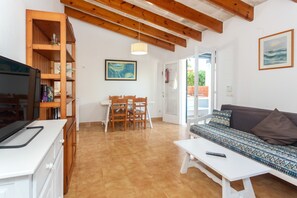 Una sala de estar con un sofá blanco, una mesa de comedor de madera con sillas, un televisor sobre un mueble blanco y una estructura de techo de madera.