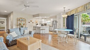 Ein geräumiges Wohnzimmer mit Sofa, Couchtisch und Essbereich mit Stühlen und Tisch. Im Hintergrund ist eine Küche mit Kühlschrank und Esstisch zu sehen.