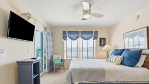 A bedroom with a bed, a TV, a chair, a ceiling fan, and a window with curtains. - Oceanfront North Myrtle Beach Condo | Balcony, Pool, Beach Access & WiFi (North Myrtle Beach)