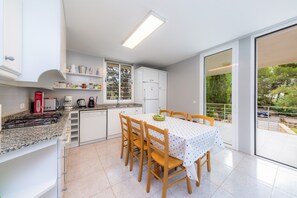 A modern kitchen with white cabinets, a dining table with chairs, and a view of greenery through a sliding glass door. - Near lake, beach and town centre - Casa Can Francesc (Alcudia)