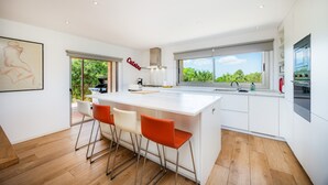 Une cuisine moderne dotée d’un grand îlot central, de tabourets de bar, d’une vue sur la verdure et d’une enseigne indiquant « Cuisine ».
