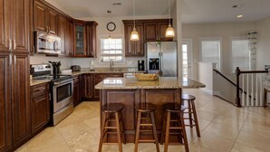 A kitchen with wooden cabinets, stainless steel appliances, a large island with stools, pendant lighting, and a view of the living area. - Oceanfront, new floors, beautiful decor, sunrise view from all decks (North Topsail Beach)