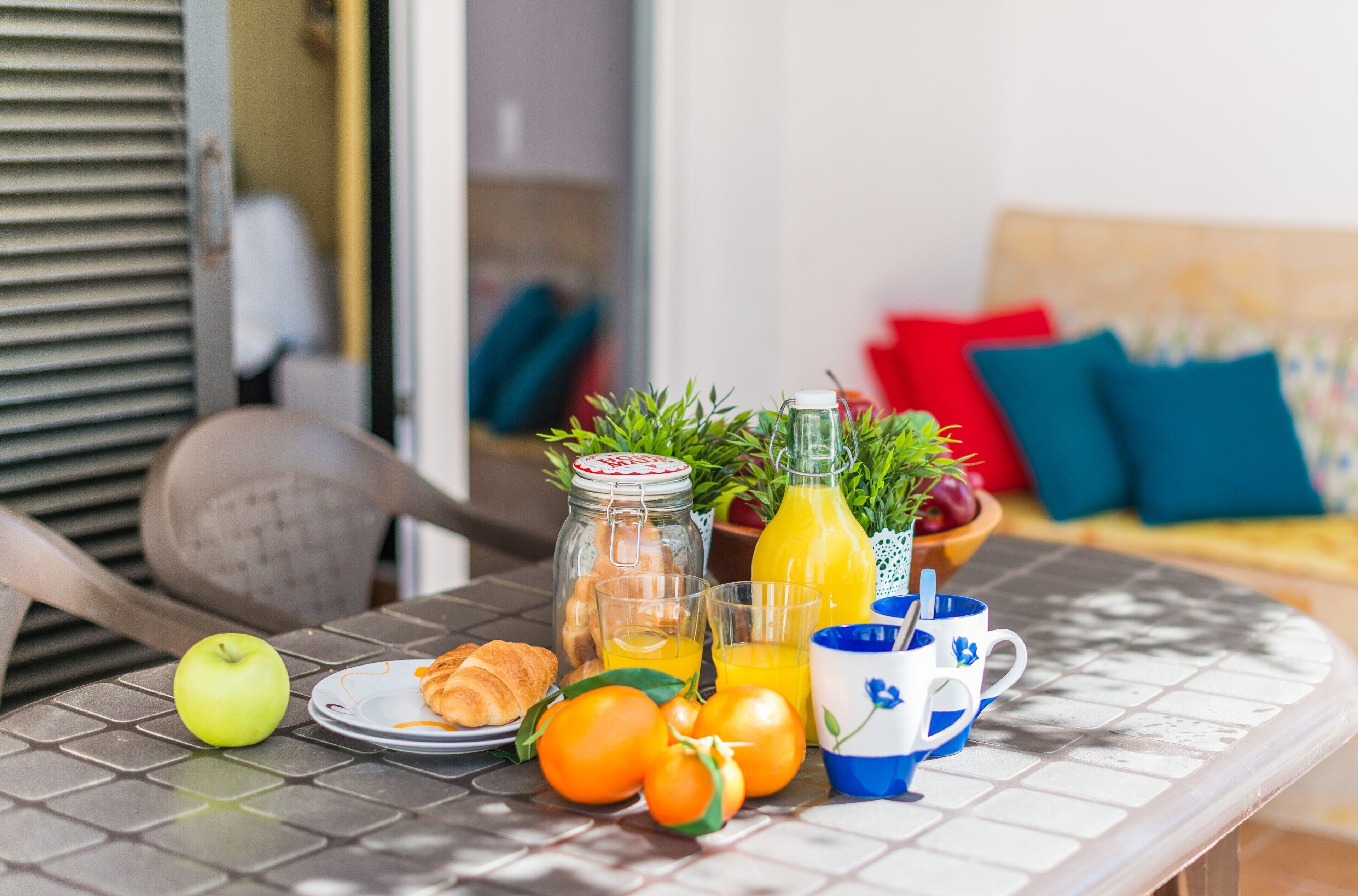 Une table de petit-déjeuner avec un bocal en verre, une bouteille de jus d’orange, une assiette de croissants, une pomme et une tasse bleue et blanche.