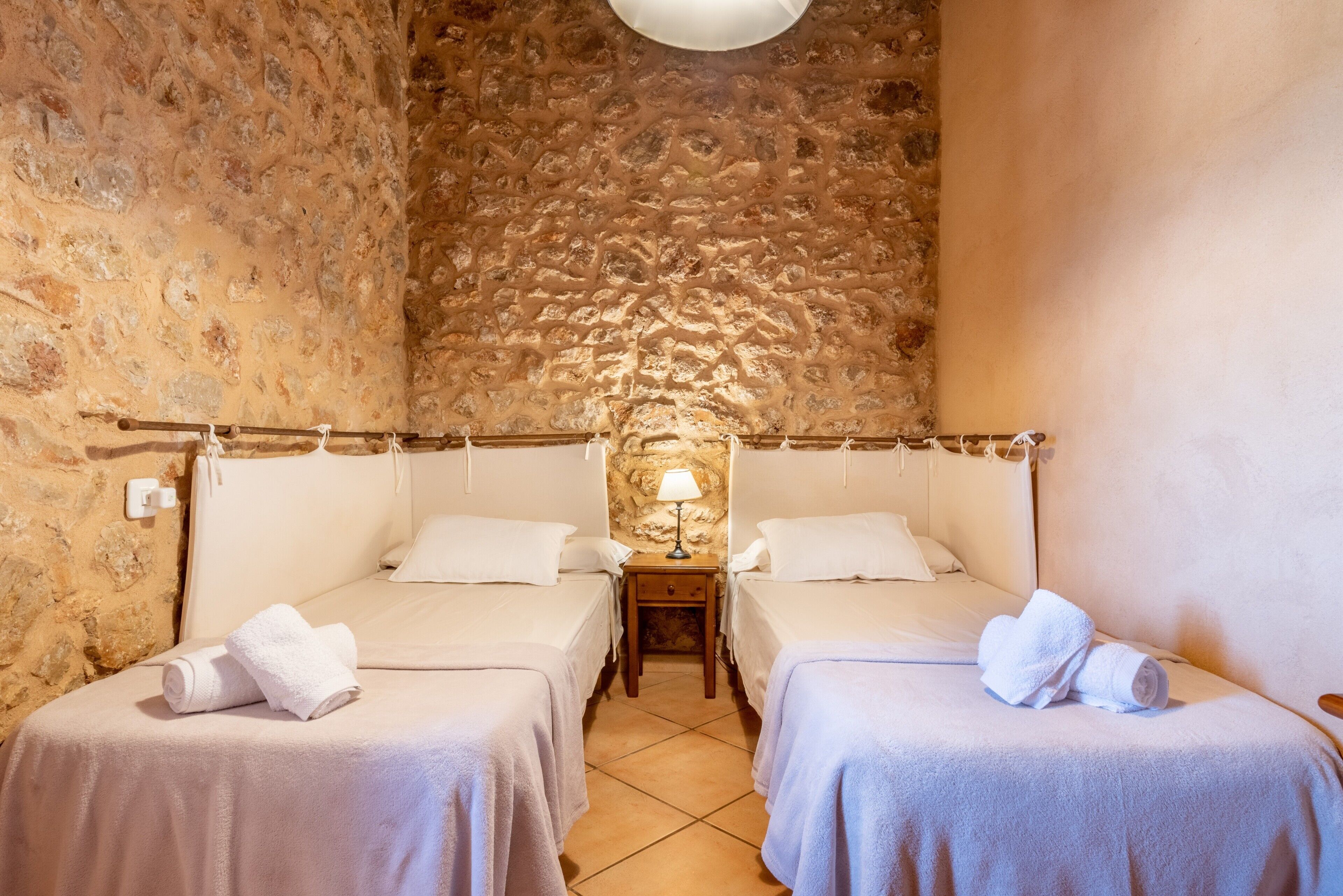 A room with two beds, a stone wall, a nightstand with a lamp, and a tiled floor.