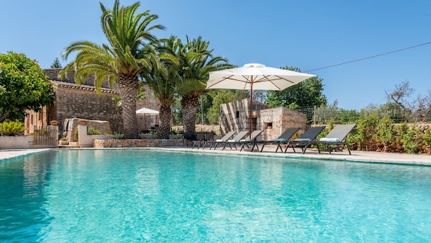 Ein klares Schwimmbecken mit Liegestühlen und Sonnenschirmen, umgeben von Steinbauten und Palmen.