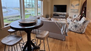 A spacious living room with a large window, a dining area with stools, a sofa, and a television. - Breathtaking views of the Ocean, Incredible Sunrise, Sunsets and Jenness Beach (Rye)