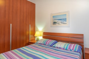 Une chambre avec une armoire en bois, un lit avec une literie à rayures, une table de chevet avec une lampe et, au mur, un tableau représentant une scène de plage.