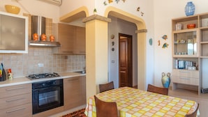 A compact kitchen with a stove, oven, and modern cabinetry. There is a dining area with a table and chairs, and a bookshelf with decorative items.