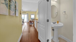 Um corredor com piso de madeira, uma porta branca, uma área de jantar com mesa e cadeiras, e um banheiro com pia e espelho.