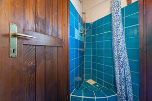 Una puerta de madera con manija metálica y cerradura, una ducha con azulejos de mosaico azules y una cortina de baño con estampado azul y blanco.