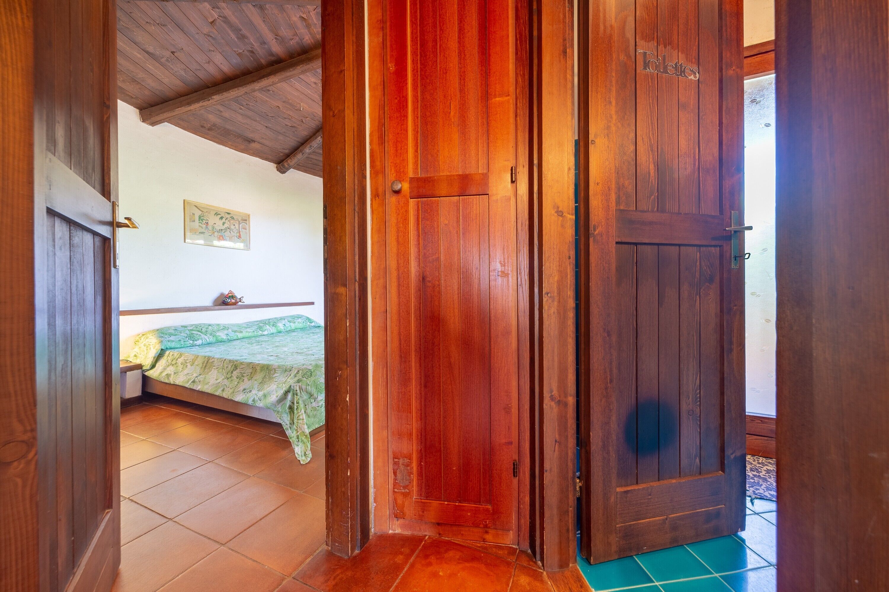 Une porte en bois avec une pancarte indiquant « Toilettes », qui donne sur une pièce contenant un lit et un couvre-lit vert.