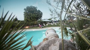 Piscina com estrutura de pedra em primeiro plano, cercada por vegetação e área delimitada com espreguiçadeiras.
