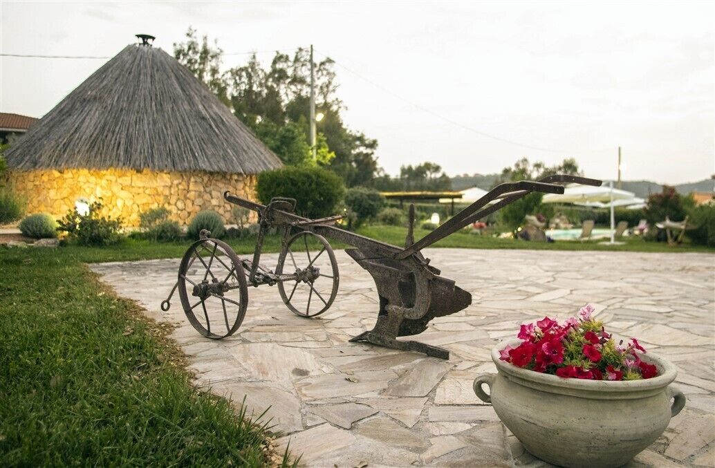 A stone pathway leading to a rustic thatched-roof structure, a vintage plow, and a flower pot.