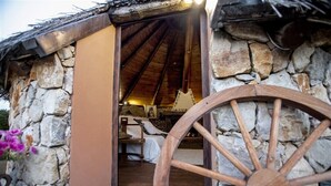 Um edifício rústico de pedra com uma roda de madeira em primeiro plano, telhado de colmo e uma cama com lençol branco.
