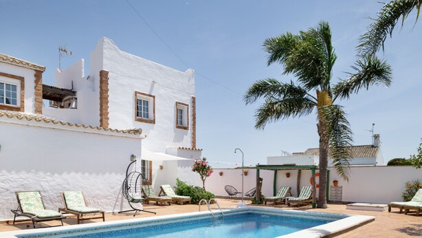 A pool area with lounge chairs, a palm tree, and a white building with windows. - Enjoy Conil Near the Sea with Pool and Barbecue – Apartment A at Villa Mayo (Conil de la Frontera)