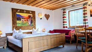 Ein Zimmer mit einem hölzernen Bett, einem roten Sofa, einem Esstisch mit Stühlen, einem Fenster mit Vorhängen und Wanddekorationen, darunter ein gerahmtes Bild und eine herzförmige Dekoration.