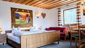 A room with a wooden bed, a red sofa, a dining table with chairs, a window with curtains, and wall decorations including a framed picture and a heart-shaped decoration.