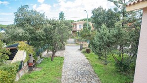 Un chemin de pierres mène à une maison dont le toit est recouvert de tuiles, et qui est entourée de verdure et d’arbres.