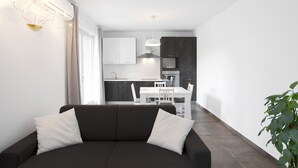 A modern living room with a dark sofa, a dining table, and a kitchen in the background. - Modern apartment near Lake Garda with Wi-Fi and terrace (BARDOLINO)