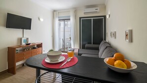 A living room with a TV, a dining table with a bowl and a glass of orange juice, a sofa, and a wall-mounted air conditioner. - Sunny Albufeira Beach Studio with Wi-Fi, Balcony, and Close to the Beach (Albufeira)