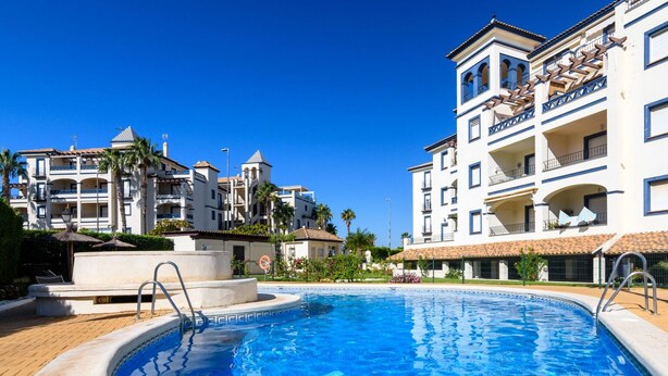 A swimming pool surrounded by multi-story buildings with balconies, palm trees, and a clear blue sky. - El Faro 36 - Two Bedroom Apartment, Sleeps 5 (Punta del moral)