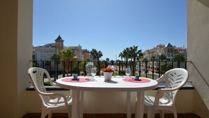 A balcony with a round table and two white chairs, overlooking a coastal area with buildings and palm trees. - El Faro 36 - Two Bedroom Apartment, Sleeps 5 (Punta del moral)