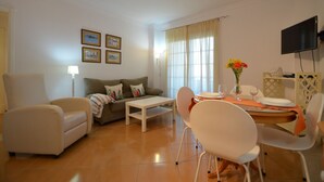 A living room with a sofa, armchairs, a coffee table, and a dining area with a table and chairs. There are framed pictures on the wall, a TV mounted on the wall, and a window with curtains. - El Faro 36 - Two Bedroom Apartment, Sleeps 5 (Punta del moral)