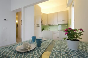 A compact kitchen with white cabinets, a dining table with a patterned tablecloth, and a potted plant with pink flowers.