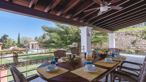 Ein überdachter Essbereich im Freien mit einem für das Frühstück gedeckten Tisch, Stühlen, einem Deckenventilator und Blick auf Garten und Pool.