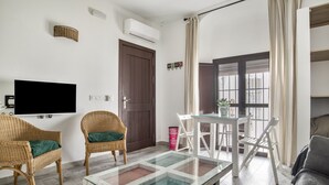 Un salon meublé avec des meubles en osier, une table basse avec un plateau en verre, un téléviseur à écran plat, une porte et une fenêtre avec des rideaux.