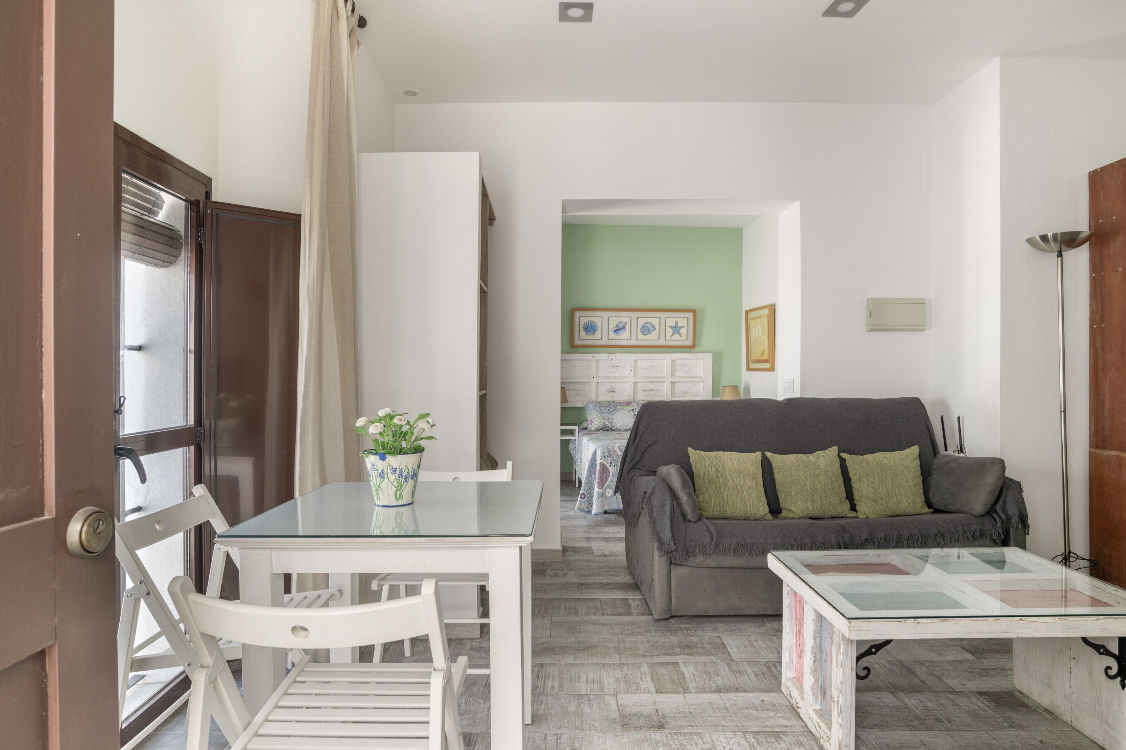 Ein Wohnzimmer mit Glastisch, weißen Stühlen, einem grauen Sofa mit grünen Kissen und einem Couchtisch.
