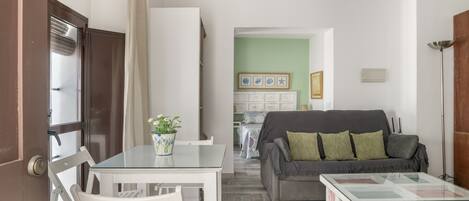 A living room with a glass table, white chairs, a grey sofa with green cushions, and a coffee table.