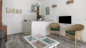 Un espace de vie compact comprenant un coin cuisine, une table à plateau en verre, des chaises en osier, un téléviseur à écran plat et des œuvres d’art murales.