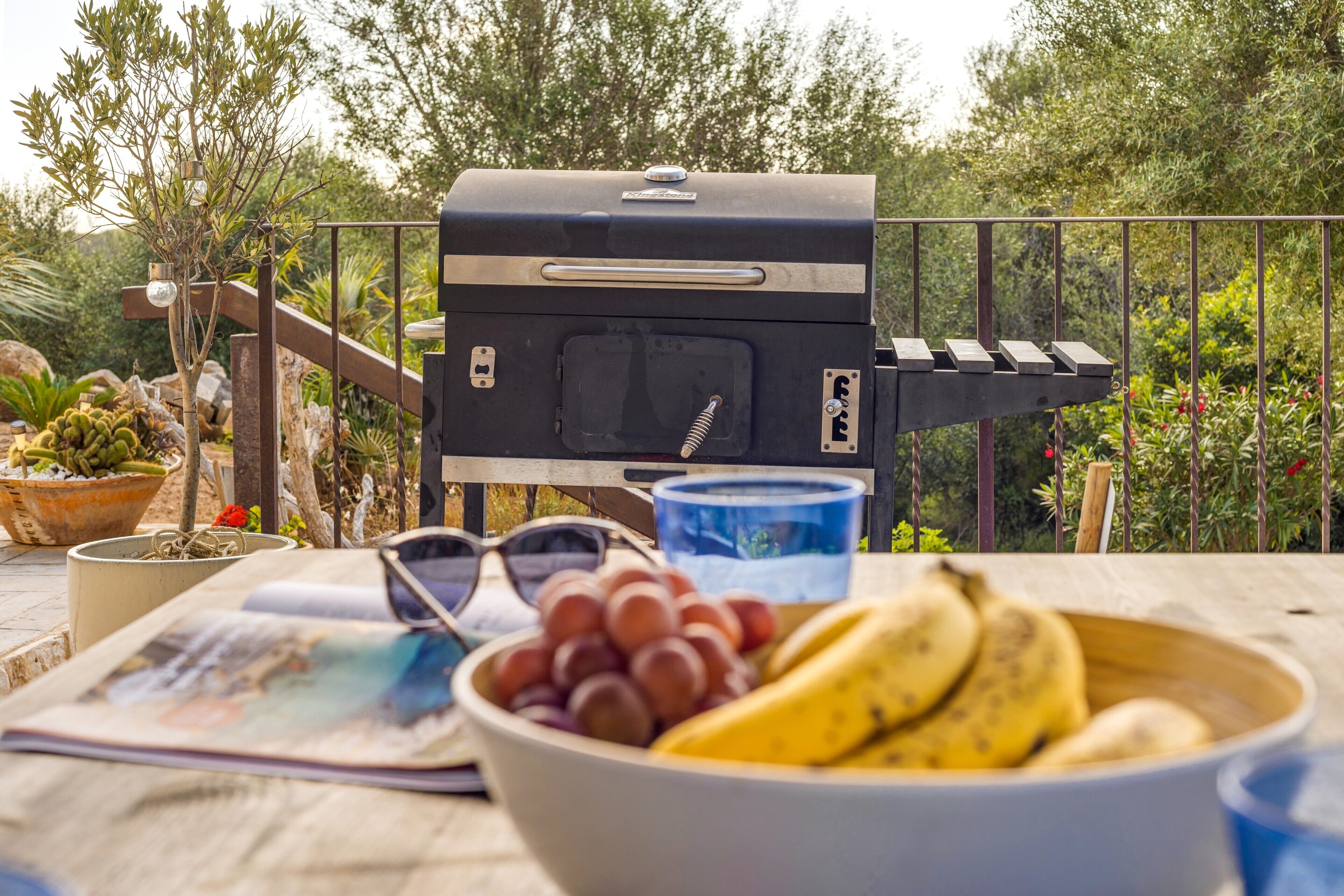 Un barbecue, un saladier de fruits, une paire de lunettes de soleil et un magazine sur une table à l’extérieur.
