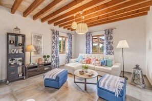 Ein Wohnzimmer mit Holzboden, einem Sofa mit gemusterten Kissen, einem blauen Hocker, einem Couchtisch und einer Kommode mit Dekoration.