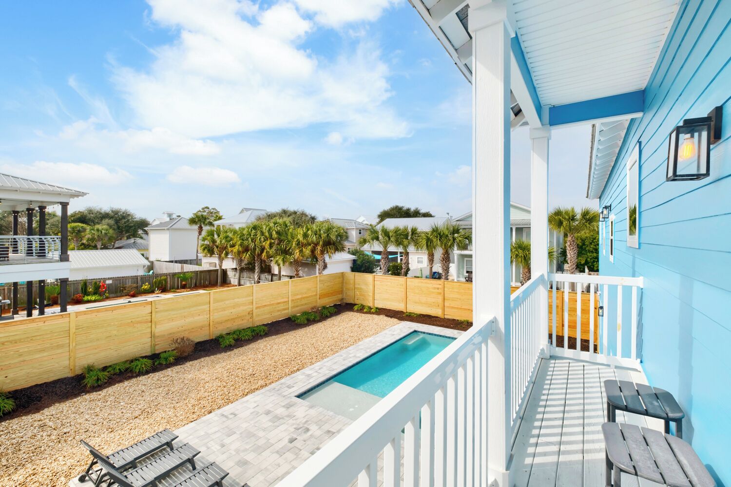 A pool area with a wooden deck, a white railing, and a view of neighboring houses and palm trees.