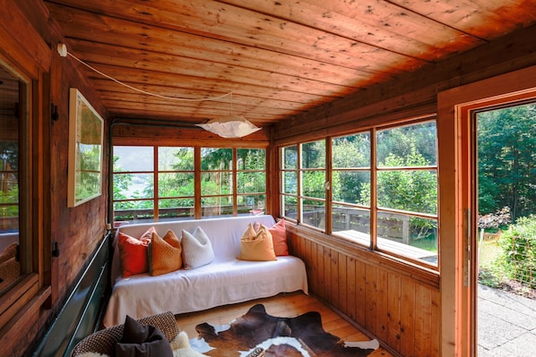 Interior de una cabaña con cama, ventanas amplias, paredes de madera y ventilador de techo.