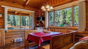 Interior de una cabaña de madera con una mesa de comedor para dos personas, ventanas amplias que ofrecen vistas a un bosque, y un techo con vigas de madera y una lámpara colgante.