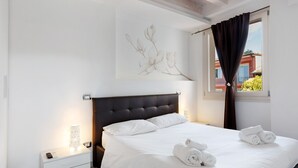 A bedroom with a large bed, a nightstand with a lamp, a wall-mounted artwork, and a window with curtains. - The Magnolia Three-Room Apartment: Charm on Three Levels in Lonato del Garda’s Courtyard (Lonato del Garda)