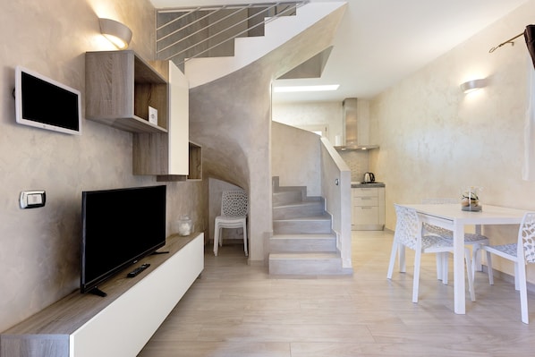 A modern interior with a flat-screen TV, built-in shelves, a staircase, a dining area with patterned chairs, and a kitchen in the background.