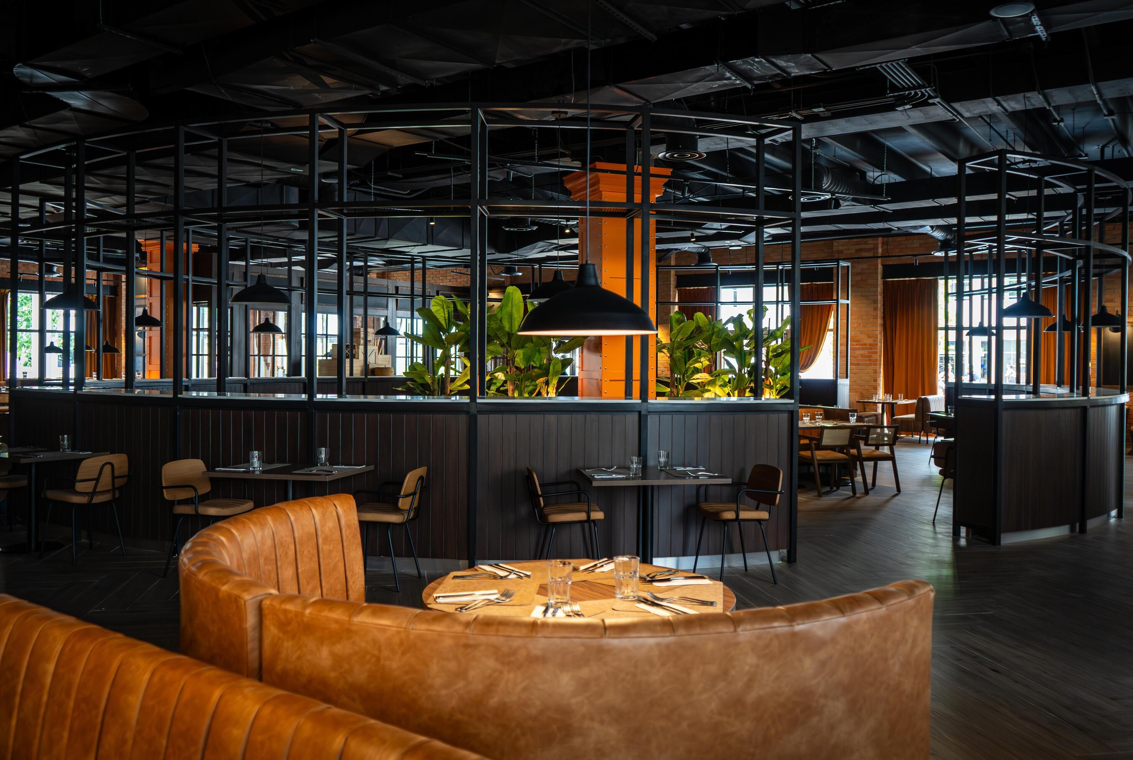 Restaurante moderno com pé direito alto, dutos aparentes e diversos tipos de assentos, incluindo boxes e mesas.
