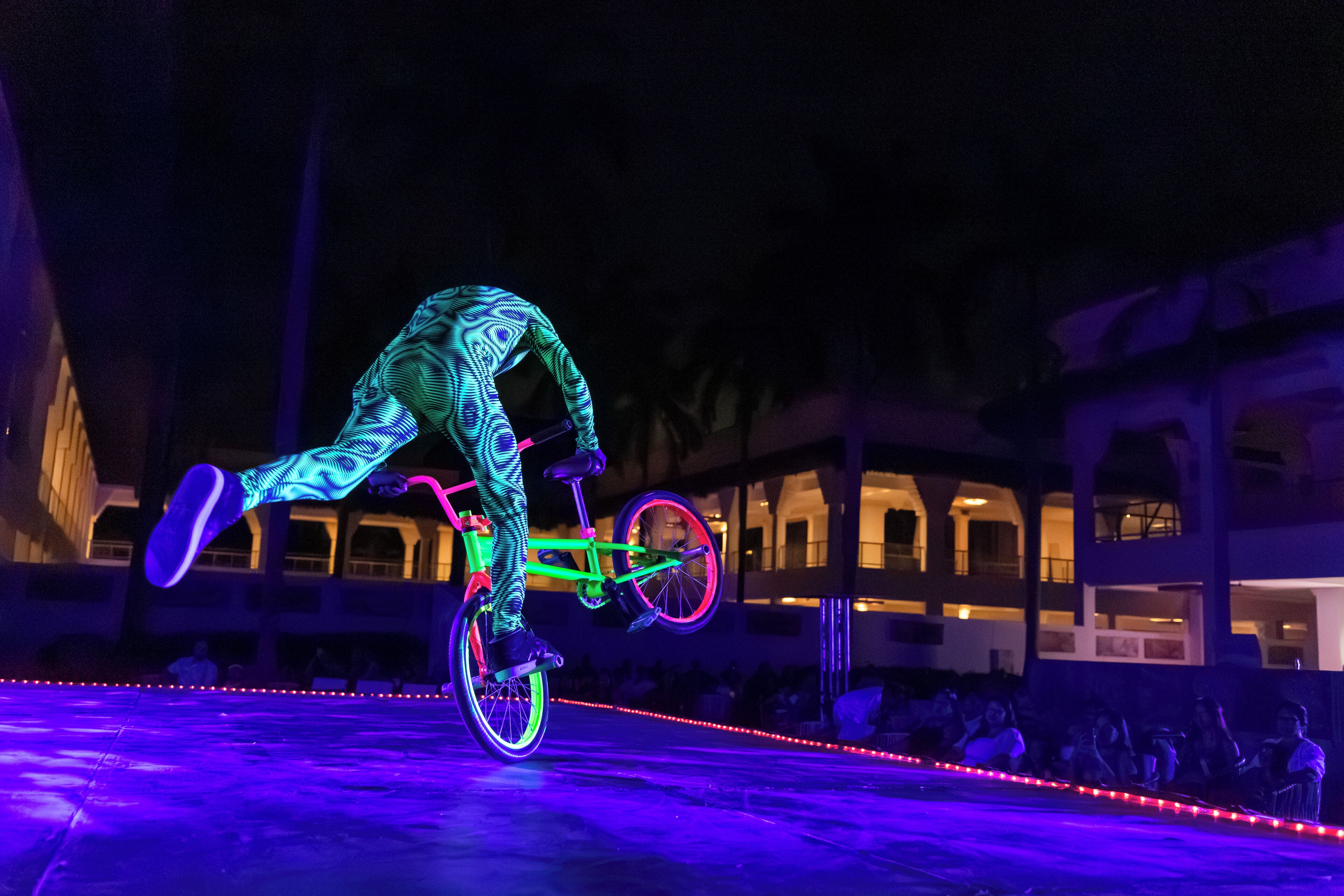 A person performing a bicycle trick on a stage with an audience in the background.
