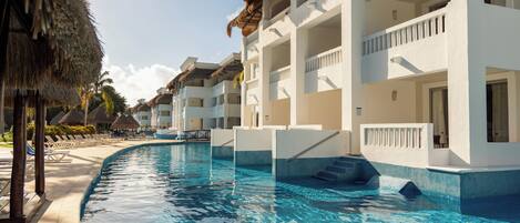 Una piscina con agua azul cristalina, rodeada de edificios blancos con balcones y una estructura de techo de paja.