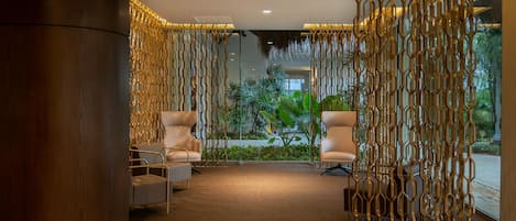 A modern lobby with a curved wooden column, two white armchairs, a patterned wall, and a view of greenery outside.