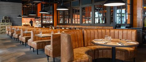 A modern restaurant interior with leather booths, round tables, pendant lighting, and large windows.