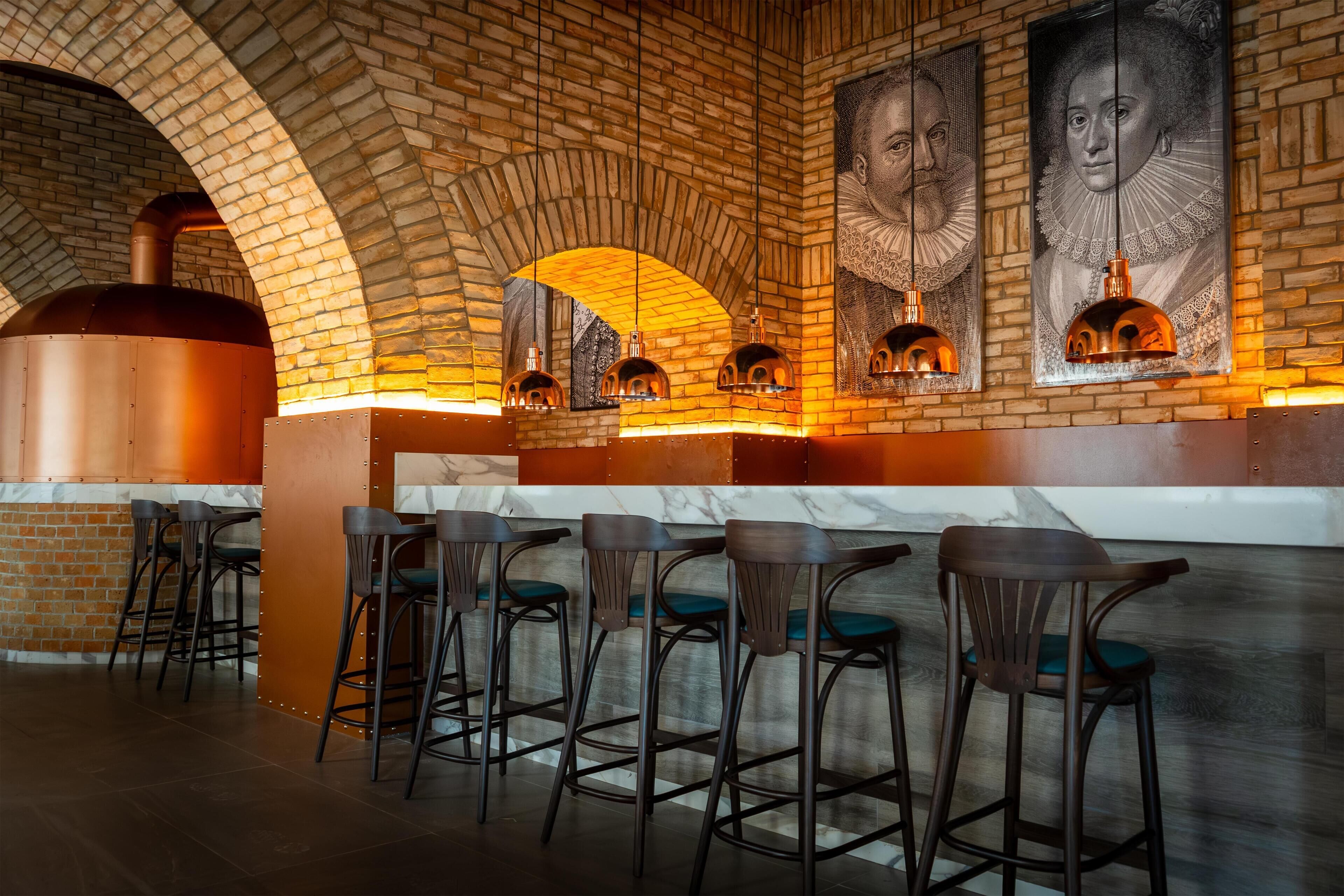 A brick-walled bar area with a large oven, bar stools, and framed portraits on the wall.