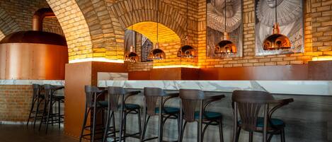 A brick-walled bar area with a large oven, bar stools, and framed portraits on the wall.