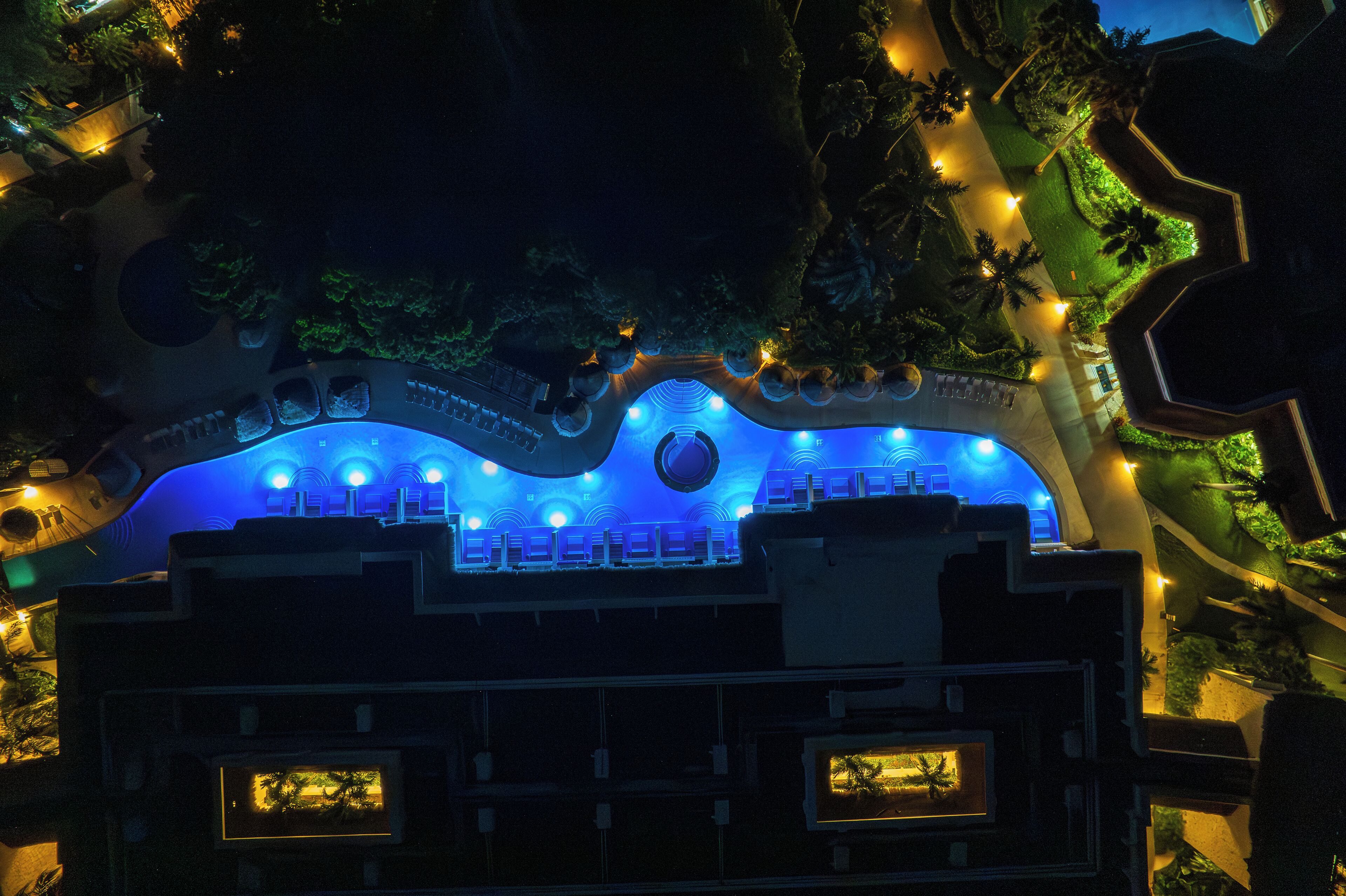 An aerial view of a hotel with a distinctive wave-like roof design, illuminated blue lights, and a landscaped garden with palm trees.